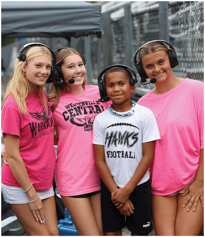 Four young people wearing headsets and casual sportswear smile while standing close together near a chain-link fence, suggesting they are involved in a sports event or broadcast. Descripci n generada con IA