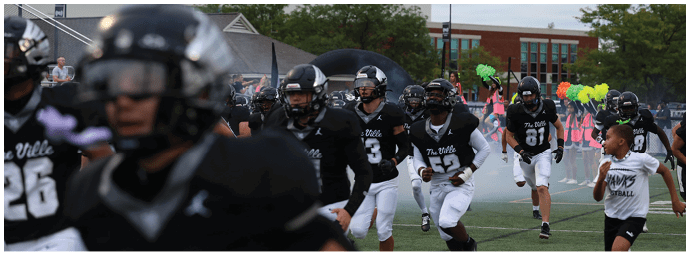 A group of people wearing black football uniforms and helmets are gathered on a field with some holding green and yellow pom-poms in the background. Descripci n generada con IA