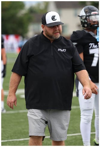 A person wearing a black “HAWKS" shirt, gray shorts, and a black and white cap is walking on a sports field with another person in a football uniform standing nearby. Descripci n generada con IA