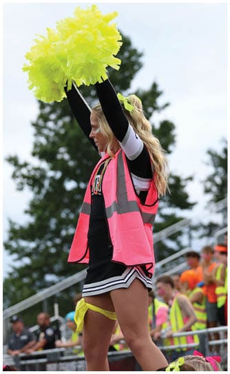 An adult wearing a cheerleading outfit and a bright pink safety vest is holding yellow pom-poms raised above their head while standing in front of a crowd at an outdoor event. Descripci n generada con IA