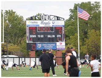 Adults in athletic clothing are stretching on a football field near a scoreboard showing a tied score and an American flag flying nearby. Descripci n generada con IA