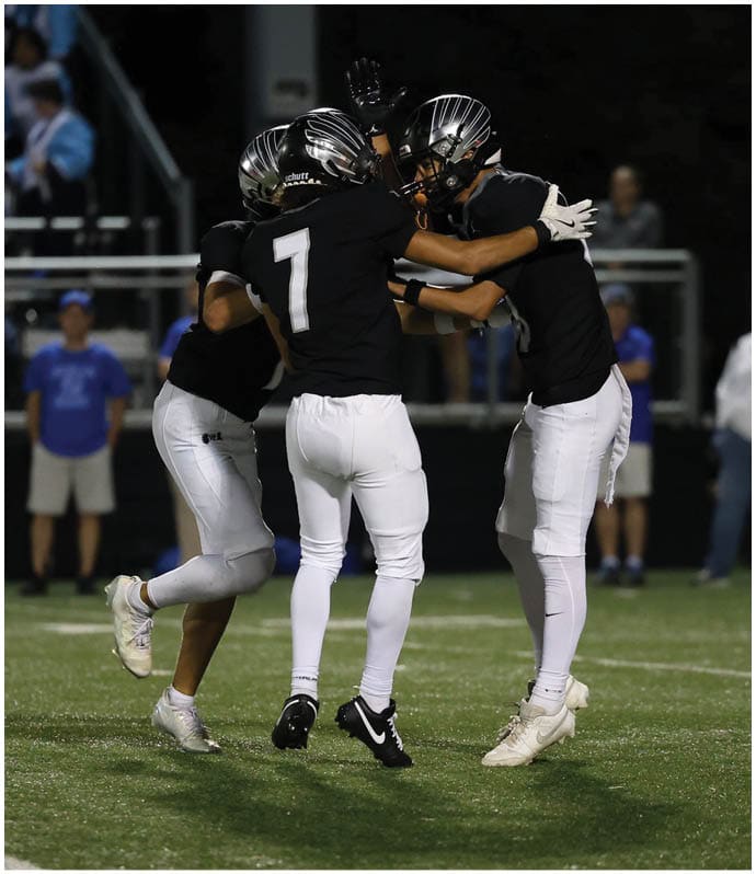 Three people wearing black football jerseys and helmets are celebrating on a football field at night. Descripci n generada con IA