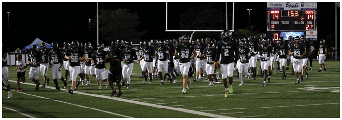 A group of people wearing black helmets and black and white sports uniforms are running on a football field at night with a scoreboard visible in the background. Descripci n generada con IA