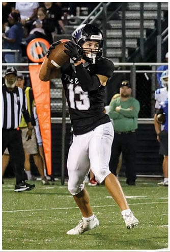 A person in a black football uniform is preparing to throw a football on a field with spectators and other players in white and blue uniforms standing in the background. Descripci n generada con IA
