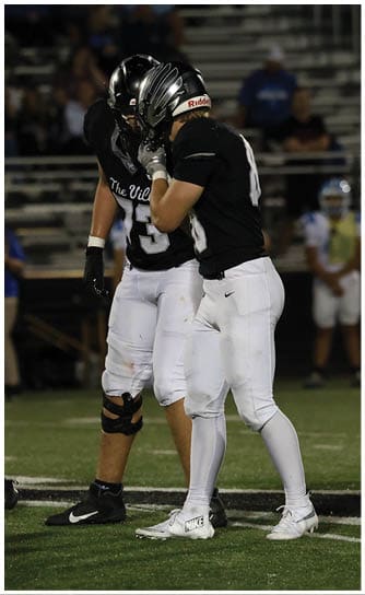Two people wearing black football helmets and black and white uniforms stand close together on a football field, with one person appearing to whisper to the other. Descripci n generada con IA