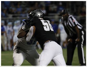 Two people in football uniforms are engaged in a play on a field with a referee observing in the background. Descripci n generada con IA