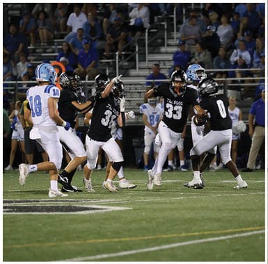 Two people in white football uniforms with “Hawks" helmets stand in the foreground while three people in green and gold football uniforms stand in the background on a field. Descripci n generada con IA