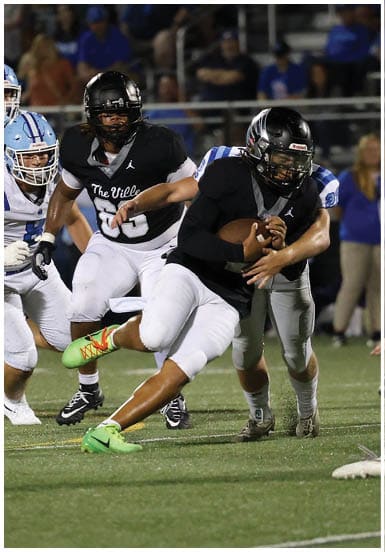 People wearing football uniforms are actively playing on a field with one person in black running with the ball while others in white and blue chase. Descripci n generada con IA