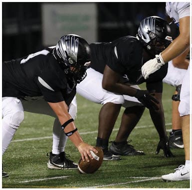 A person in a black sports jersey and white pants is crouched in a ready position on a football field wearing a black and silver helmet. Descripci n generada con IA