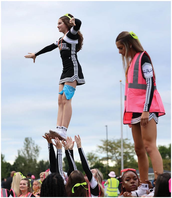 A person in a cheerleading uniform is being lifted into the air by a group of people during an outdoor event with others watching nearby. Descripci n generada con IA