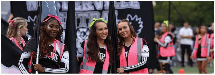 Three people wearing bright pink vests and colorful headbands hold flags and smile in front of a banner that reads “WARRVILLE." Descripci n generada con IA