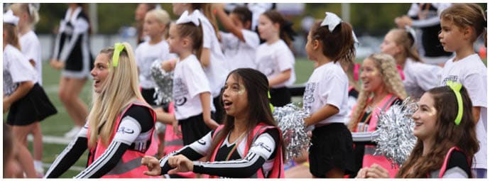 Adults and children wearing cheerleading uniforms and holding pom-poms are enthusiastically performing on a sports field. Descripci n generada con IA