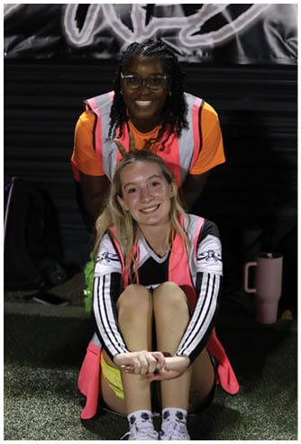 Two adults wearing bright pink safety vests are smiling, with one sitting on the ground hugging their knees and the other standing behind them on a grassy surface at night. Descripci n generada con IA