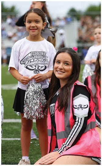 A person wearing a white shirt with “Future Warhawk Cheerleader" text holds silver pom-poms while another person with long dark hair smiles and wears a black and pink outfit. Descripci n generada con IA