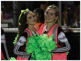 A person wearing a black and white cheerleading uniform with the word “CENTRAL" and a red, white, and blue flower lei stands in front of a blurred background with a fence and people. Descripci n generada con IA