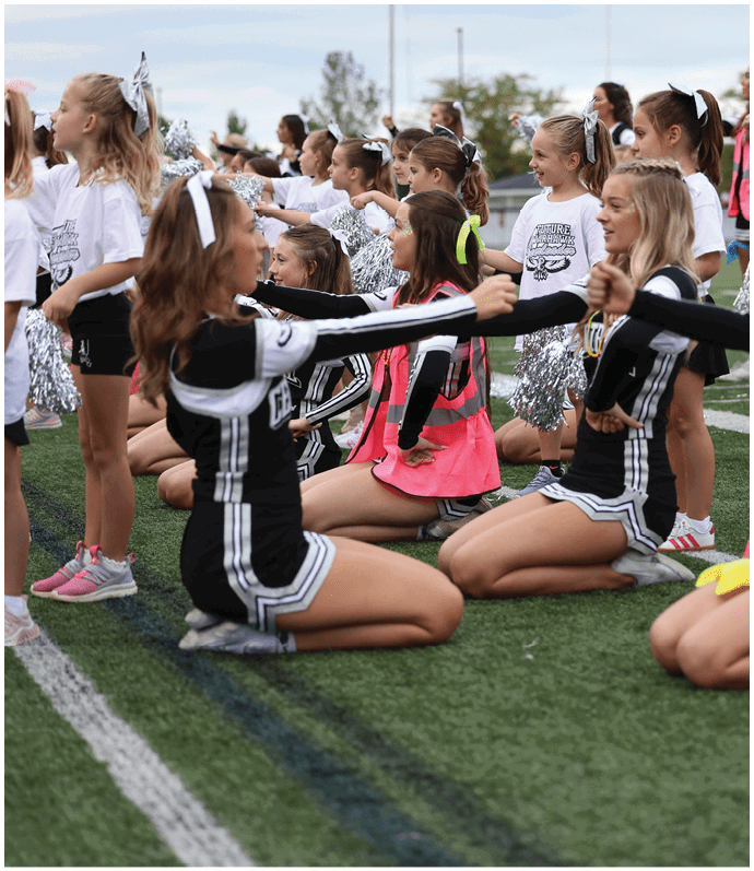 A group of people in cheerleading uniforms and bright vests are kneeling on a sports field performing a coordinated routine with pom-poms. Descripci n generada con IA