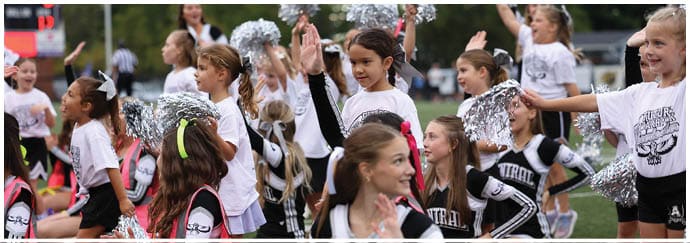 Children wearing cheerleading uniforms and holding pom-poms are gathered outdoors, some raising their hands and smiling. Descripci n generada con IA
