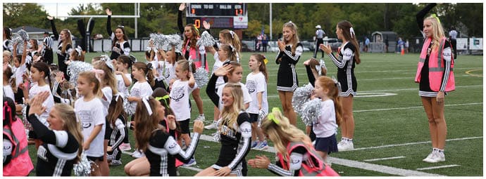 Children and adults in cheerleading uniforms are performing a routine on a sports field with some participants kneeling and others standing. Descripci n generada con IA