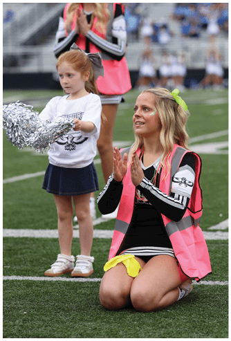 An adult wearing a pink vest and a yellow bow is kneeling on a sports field clapping while a child in a white shirt and navy skirt stands nearby holding silver pom-poms. Descripci n generada con IA