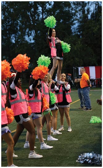 A group of people wearing cheerleading uniforms and pink safety vests perform a cheer routine on a sports field at dusk while holding green and orange pom-poms. Descripci n generada con IA