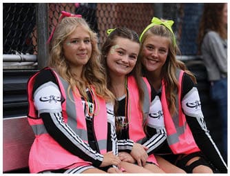 Three people wearing matching black and white outfits with bright pink safety vests and colorful bead necklaces are smiling and sitting close together. Descripci n generada con IA