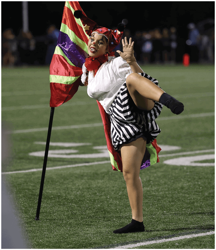 A person wearing a striped skirt and colorful costume is performing a high kick next to a flag on a sports field. Descripci n generada con IA