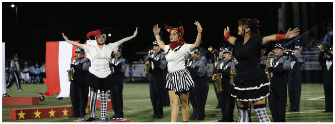Three people wearing colorful costumes and patterned tights stand on decorated podiums on a field with a band playing instruments in the background. Descripci n generada con IA