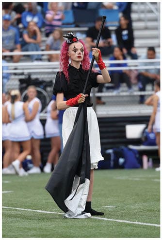 Person with curly pink hair wearing a black top, white skirt, and red gloves stands on a sports field holding a black and white flag. Descripci n generada con IA