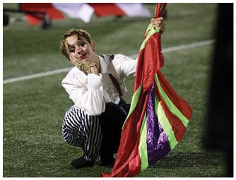 A person wearing striped pants and a white shirt is crouching on a field while holding a colorful flag. Descripci n generada con IA