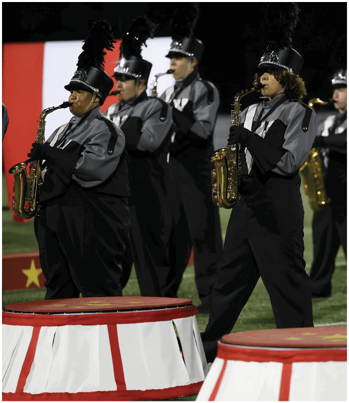 People in marching band uniforms are playing saxophones on a field at night. Descripci n generada con IA