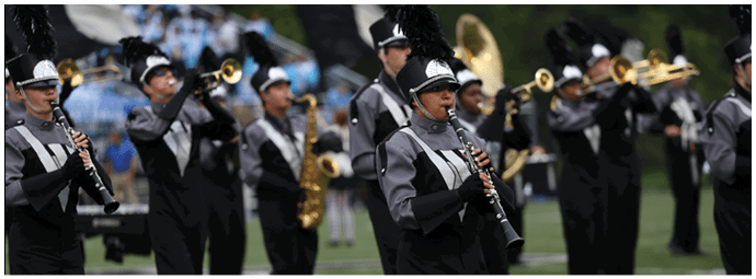 People in matching band uniforms are playing various brass and woodwind instruments outdoors during a performance. Descripci n generada con IA