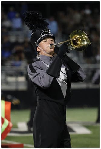 A person in a marching band uniform with a feathered hat is playing a brass instrument on a football field with colorful flags and a crowd in the background. Descripci n generada con IA