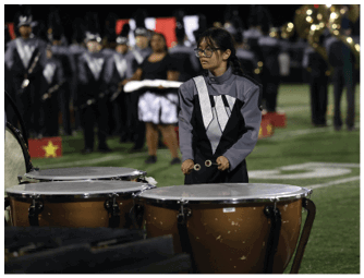 A person wearing a gray and black marching band uniform is playing timpani drums on a football field with other band members visible in the background. Descripci n generada con IA