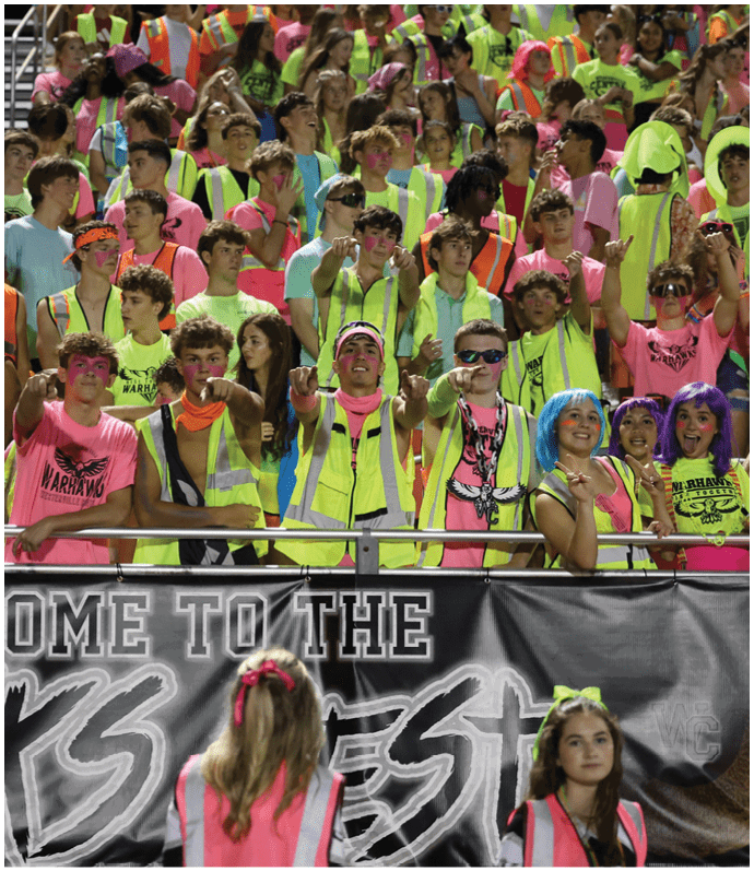A large group of people wearing brightly colored neon clothing and safety vests are gathered closely together, some with painted faces and accessories, appearing excited and energetic in a stadium setting. Descripci n generada con IA