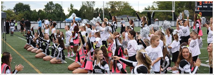A large group of people, many wearing cheerleading uniforms and holding pom-poms, are gathered on a sports field, cheering and raising their arms. Descripci n generada con IA