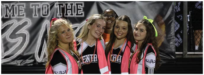 Four people wearing cheerleading uniforms and bright pink safety vests stand closely together smiling in front of a dark background. Descripci n generada con IA