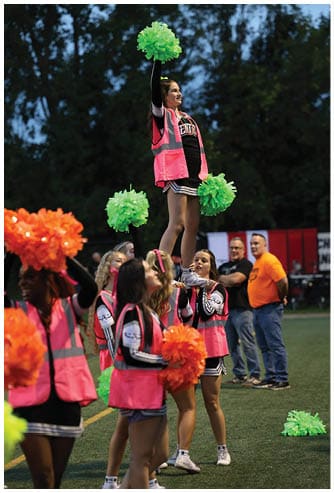 A person wearing a cheerleading outfit and a pink safety vest is holding green pom-poms, raising one arm, and smiling outdoors with trees in the background. Descripci n generada con IA