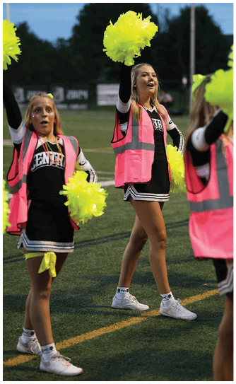 A person wearing a black and white outfit with a pink safety vest is holding bright yellow pom-poms and appears to be cheering outdoors. Descripci n generada con IA