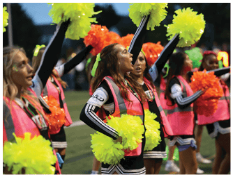 People wearing matching outfits and holding colorful pom-poms are cheering energetically on a sports field. Descripci n generada con IA