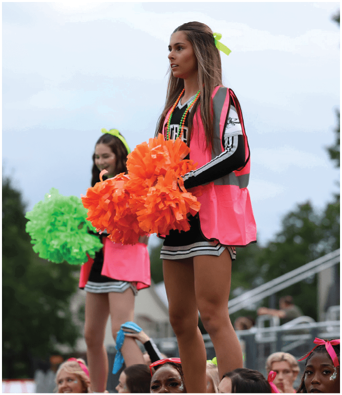 Two people wearing cheerleading outfits and neon pink vests hold colorful pom-poms while standing above a crowd at an outdoor event. Descripci n generada con IA
