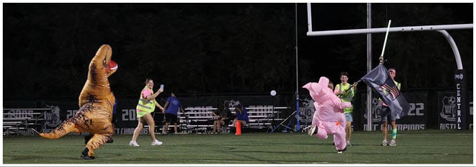 People in dinosaur and animal costumes are playing or running on a football field at night. Descripci n generada con IA