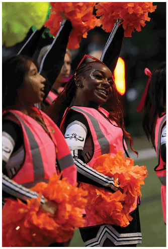 A group of people wearing cheerleading uniforms and pink vests are holding orange and green pom-poms while performing on a field. Descripci n generada con IA