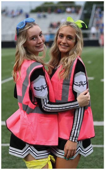 Two adults wearing cheerleading outfits and bright pink safety vests are standing on a sports field, smiling and embracing each other. Descripci n generada con IA
