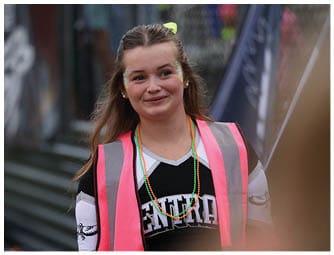 A person wearing a black and white sports jersey and a bright pink safety vest is standing outdoors near a sports field with a slight smile. Descripci n generada con IA