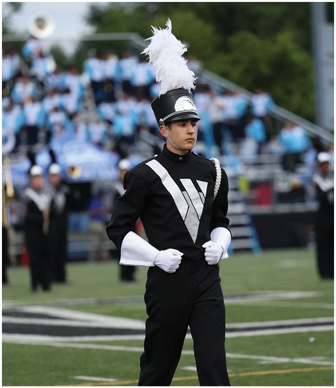 A person in a black marching band uniform with white gloves and a tall hat with a white plume stands on a football field with a blurred marching band and crowd in the background. Descripci n generada con IA