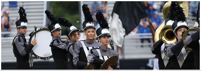 People in marching band uniforms with tall black plumes on their hats are playing cymbals and drums on a sports field with spectators in the background. Descripci n generada con IA