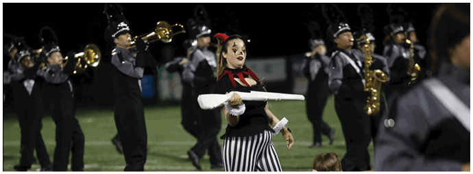 A person in striped pants and a black top is performing with a baton on a field while a marching band plays in the background. Descripci n generada con IA