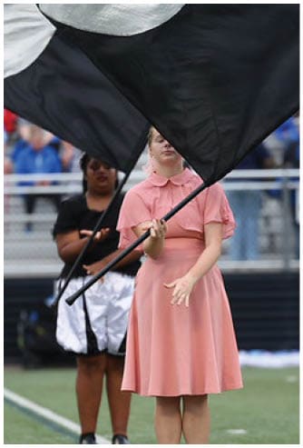 A person in a pink dress is holding and waving a large black flag on a sports field with another person in the background also holding a flag. Descripci n generada con IA
