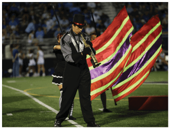 A person in a marching band uniform and top hat is playing a clarinet on a field while color guard members wave red, yellow, and purple striped flags in the background. Descripci n generada con IA