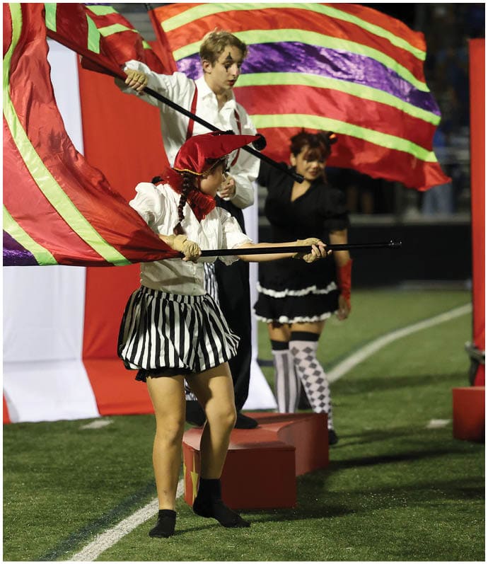 People dressed in colorful costumes perform with large striped flags on a sports field during an event. Descripci n generada con IA
